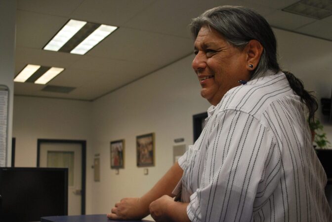 Side photo of an Indigenous man who is smiling inside an office. He is wearing his long hair in a ponytail and wears a striped button down shirt.