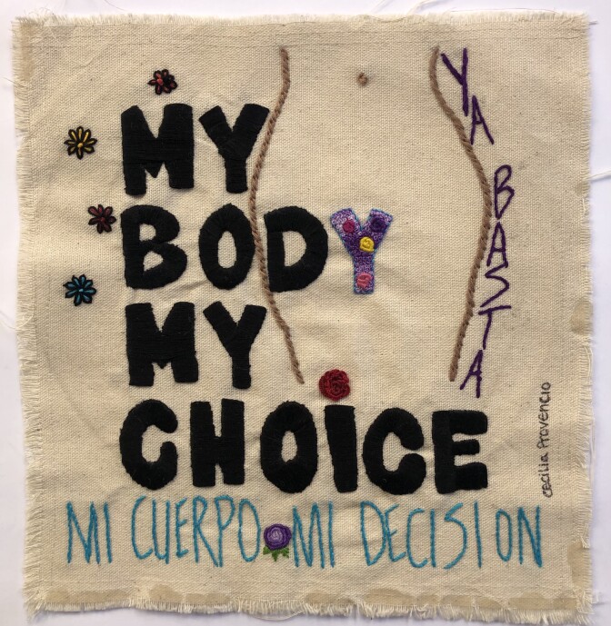 A quilt square primarily comprised of a beige background fabric. On it, the words "My body, my choice," are sitched on with a black, bold lettering. Behind it, an outline of woman's body is stitched on with a dark brown thread. The "Y" from "body" lines up with the outline, serving as the woman's crotch. Colorful flowers decorate the inside of the "Y." Along one side of the body the words, "Ya basta" are stitched on in purple thread. Underneath, "My body, my choice," are the words, "Mi cuerpo, mi decision" in blue thread with a purple flower crocheted in the middle.