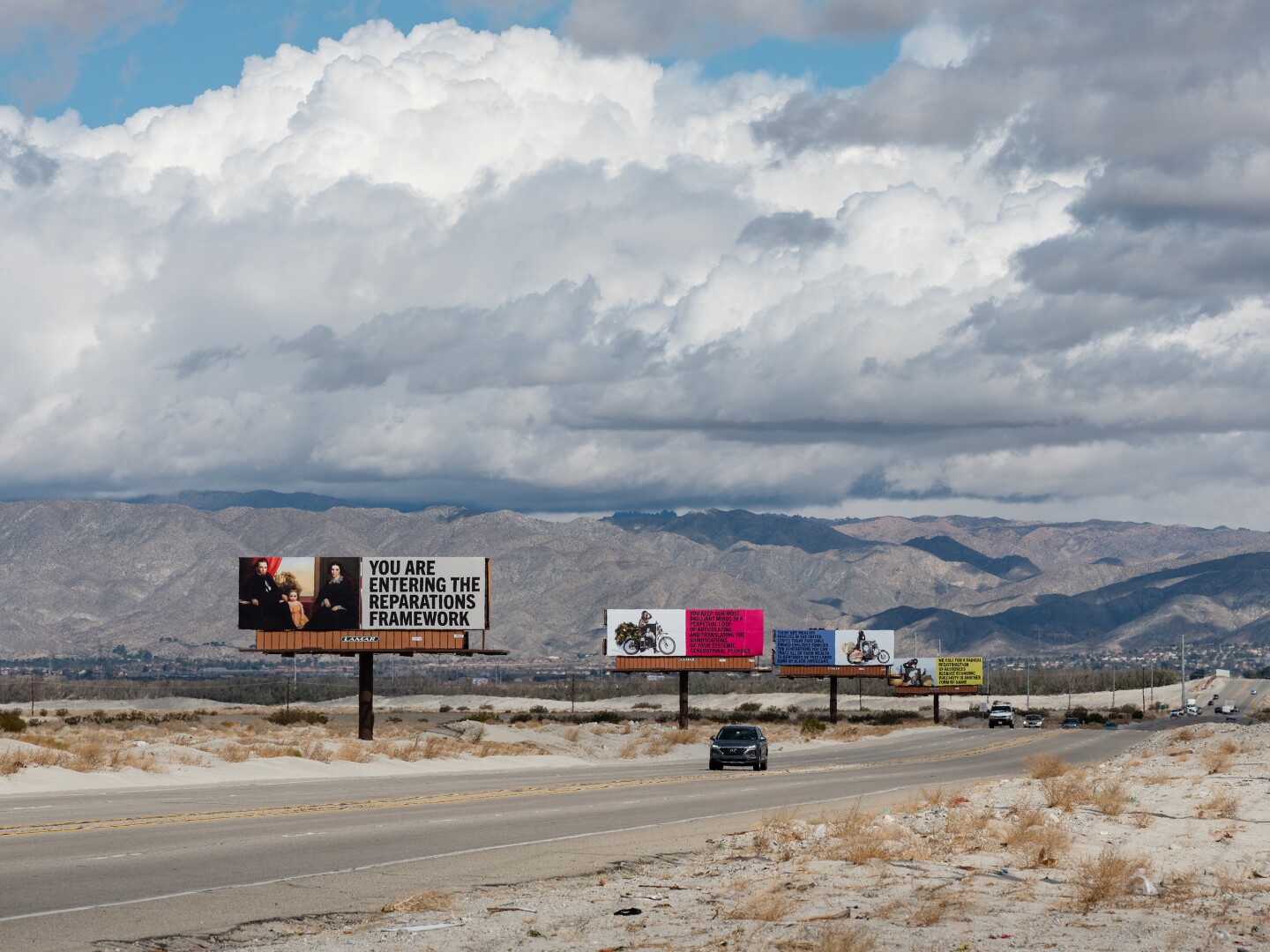 Xaviera Simmons' billboard art installation, "Because You Know Ultimately We Will Band a Militia," for Desert X 2021 stands along the Gene Autry Trail. Four billboards stand in a line. The one closest to the foreground reads, "You are entering the reparations framework." A car drives down the road that runs along the billboards.