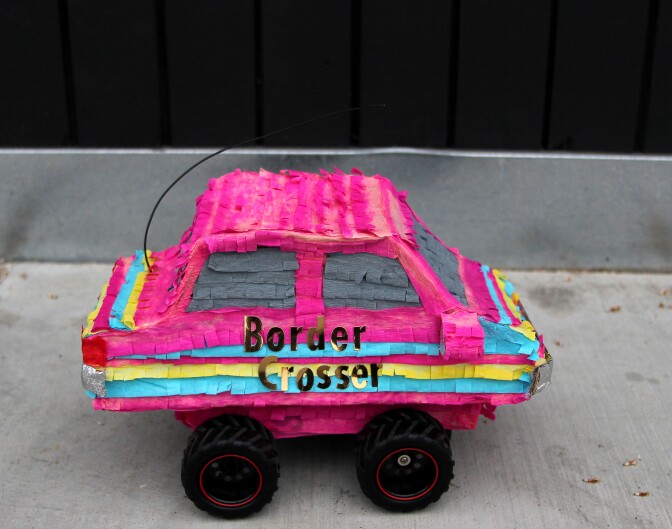 A colorful pink car striped with yellow and blue is made out of crepe paper and reads "Border Crosser."