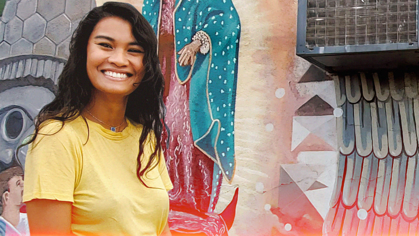 A woman in a yellow shirt smiles in front of a wall painting of Virgen de Guadalupe.