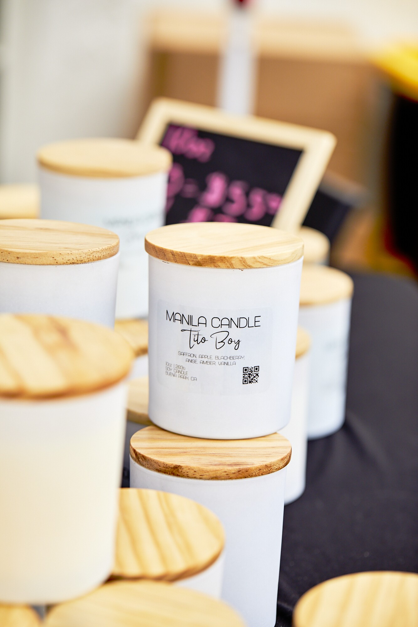 The Tito Boy candle sits at the Manila Candle booth at the FilLed Market in Downtown L.A. The candle body is white and reads, "Manila Candle, Tito Boy" in black print. The candle lid is a light-colored wood.