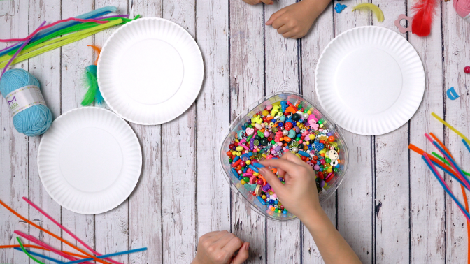 A table with paper plates, pipe cleaners, yarn, a container full of colorful beads and feathers all laid out.
