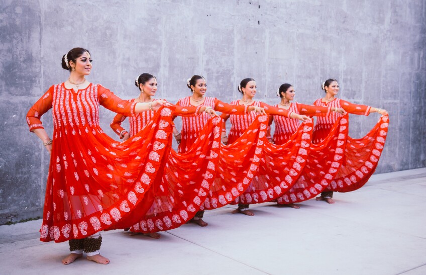 Six women of South Asian descent stand in a diagonal line, wearing long, red long-sleeved dresses with white embroidered detailing. Each woman is holding up the left side of their skirt, creating a half-circle shape down the row.