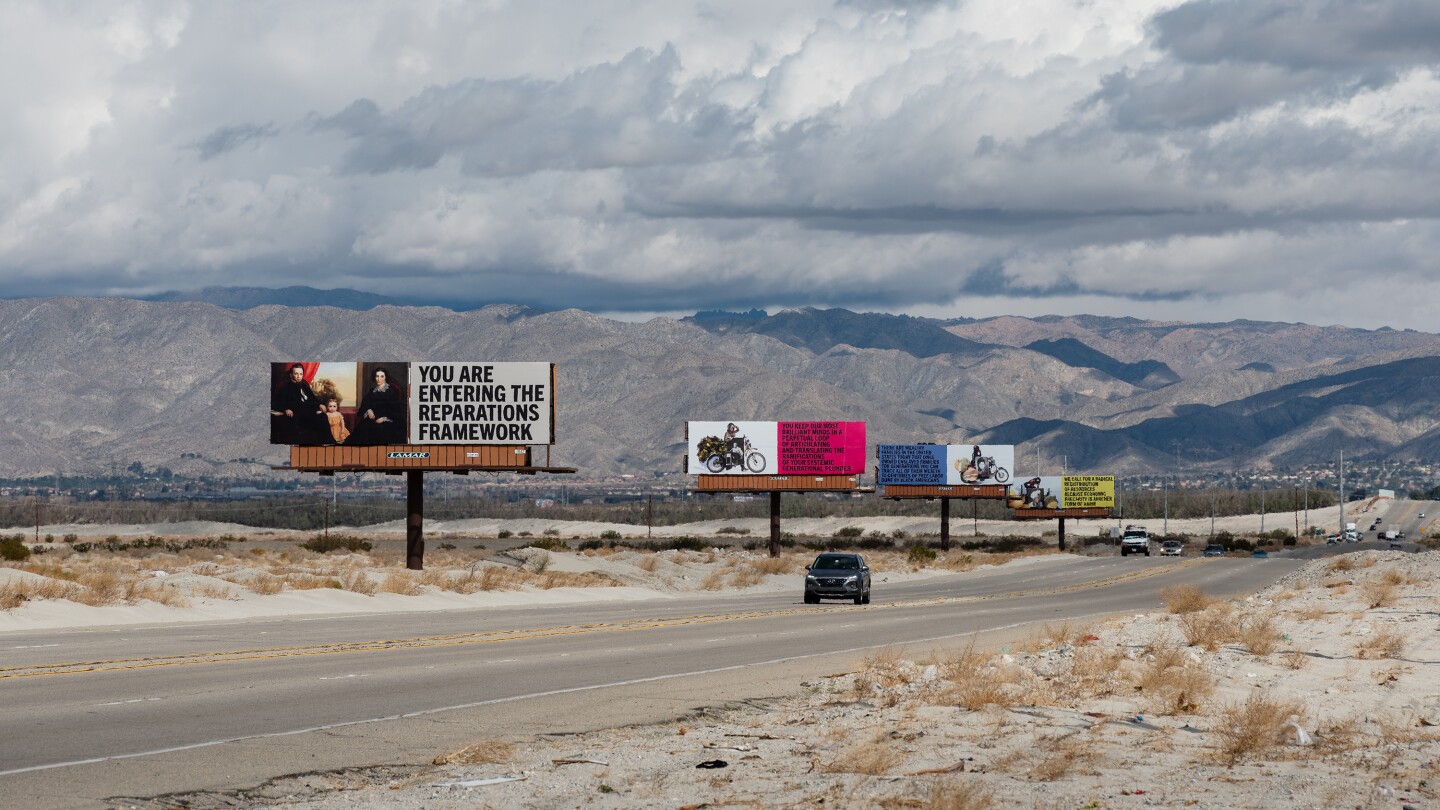 Xaviera Simmons' billboard art installation, "Because You Know Ultimately We Will Band a Militia," for Desert X 2021 stands along the Gene Autry Trail. Four billboards stand in a line. The one closest to the foreground reads, "You are entering the reparations framework." A car drives down the road that runs along the billboards.