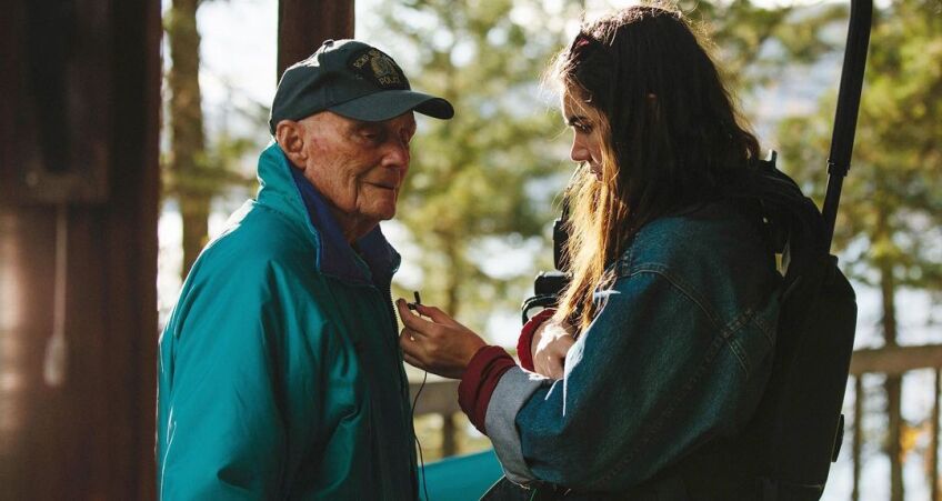 An elderly man in a teal green-blue windbreaker stands still as a younger woman in a denim jacket clips a lavalier mic to his jacket. The woman is equip with a camera rig on her back.