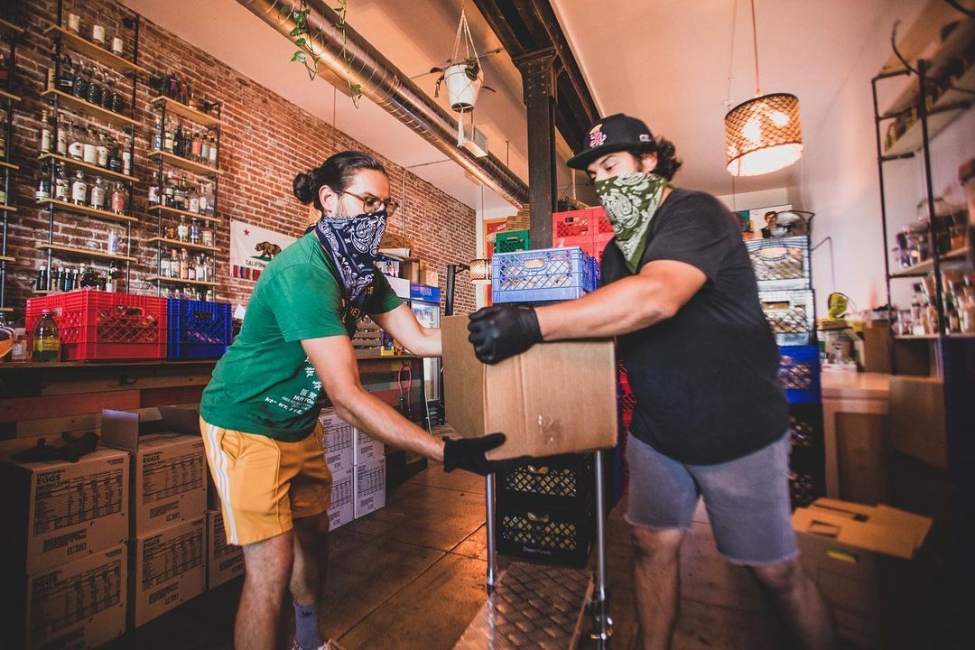 A volunteer at No Us Without U hands a cardboard box of produce to another volunteer. They are indoors in a restaurant that is being used as a hub for food distribution. Both of the volunteers have handkerchiefs tied over their mouths to serve as face masks and they are both wearing black rubber gloves.