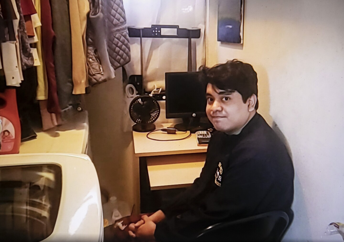 A young man raises his eyebrows at the camera as he sits at a cramped desk next to a washing machine and clothes hanging on a rack.