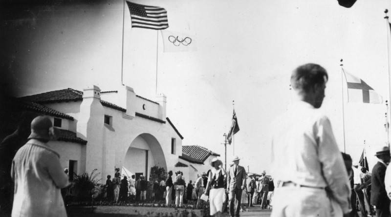Men and women gather around under flags and the main entrance and headquarters of the Olympic Village in Baldwin Hills during the 1932 Olympics in Los Angeles.