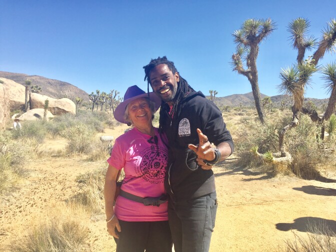 Eva Zoltes, left, is wearing a bright pink t-shirt and has a holster strapped around her hips, carrying a sound recording device. A small microphone and chords are clipped on her shirt. She is wearing a purple, wide-brimmed hat as she poses with Kennedy Verrett, left. Kennedy is wearing a black zip-up hoodie with a design over the right breast that reads, "Harrison House." Kennedy is smiling wide as he holds his hand up, waving for the photo.
