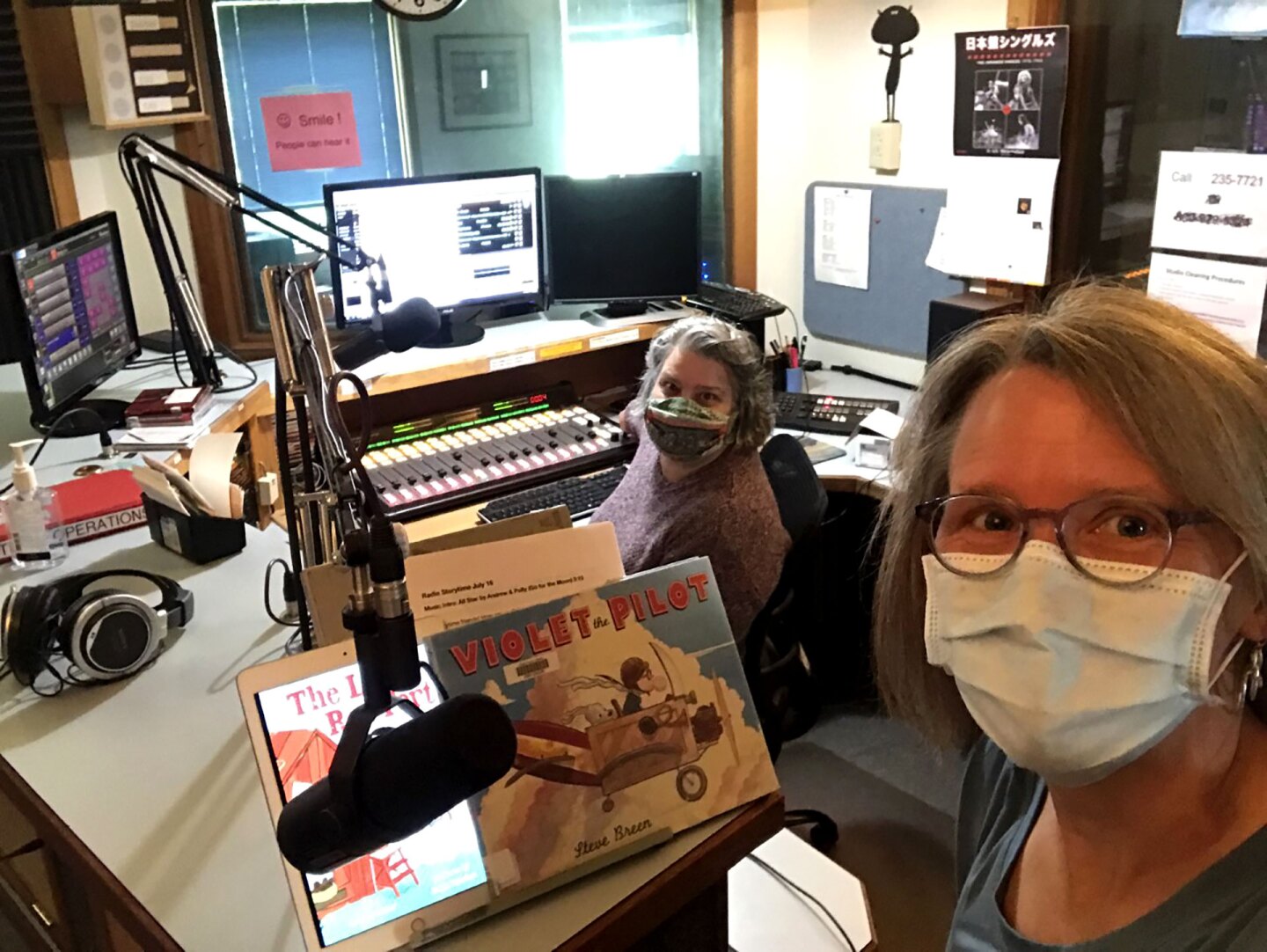 Two women wearing masks sit inside a radio recording studio