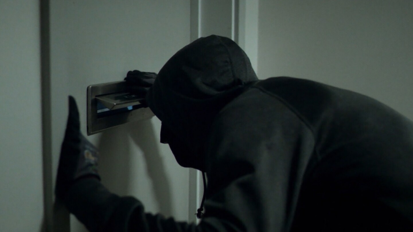 A man wearing dark clothes looks through a door's mail slot. | Still from "Follow the Money" Season 2, Episode 5