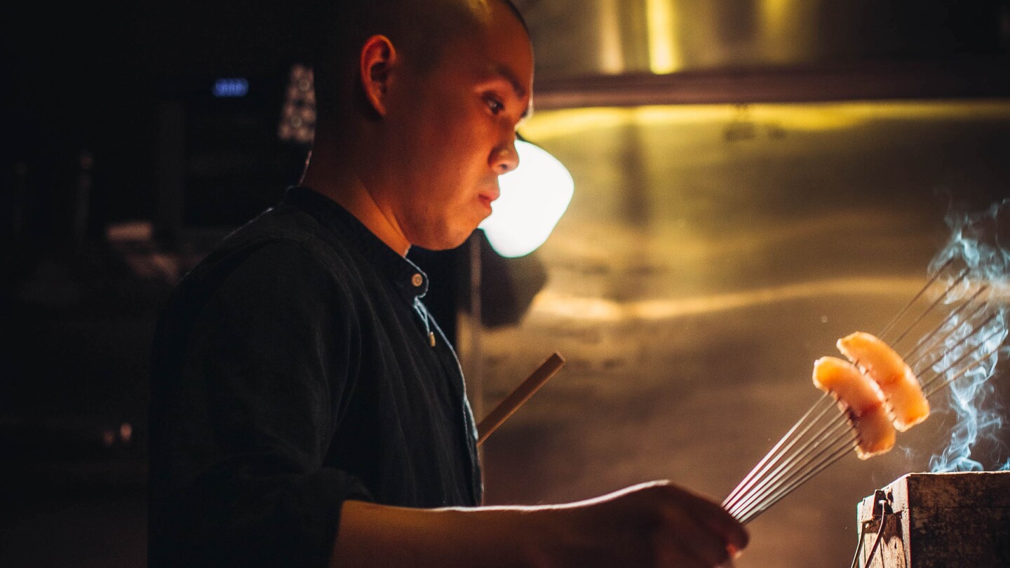 Chef Jon Yao cooks two skewered, bite-sized pieces of fish over a smokey box. He's standing in a dark kitchen and his face is lit by a warm, orange light coming from beneath him.
