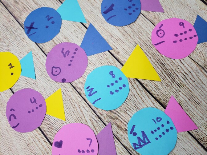 Colorful cutouts shaped like fish, each labelled with a number from one to 10, are lined up on a wooden table.