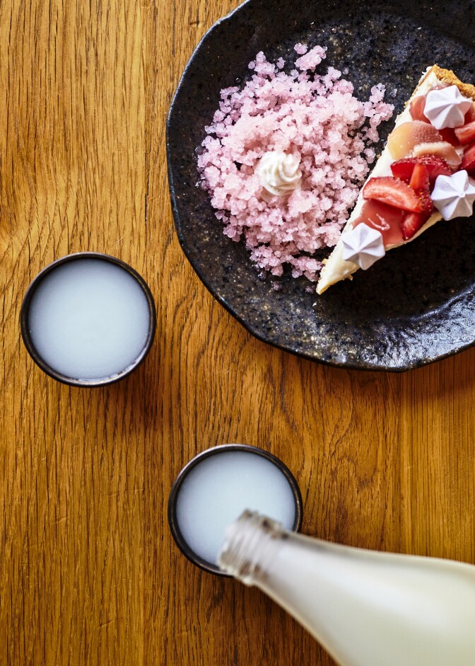 Cloudy nigori is poured in a cup beside a plate of ume cheesecake topped with strawberry rhubarb jellies, hibiscus meringue and red shiso granita.