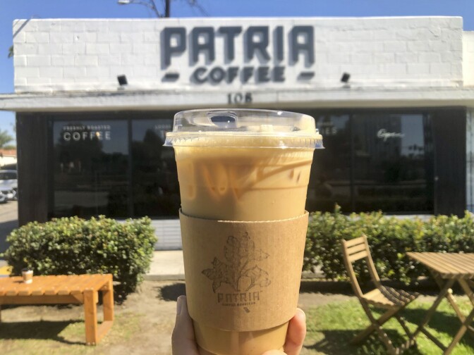 A hand holds up a cup of iced coffee in a plastic cup. Behind the cup of coffee is a building that reads, "Patria Coffee."