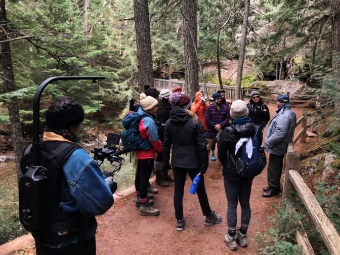 A woman equip with a camera rig captures footage of a group of hikers in the middle of a trail. The group of hikers are huddled around an elderly man wearing a puffer jacket. Around the trail are towering trees.