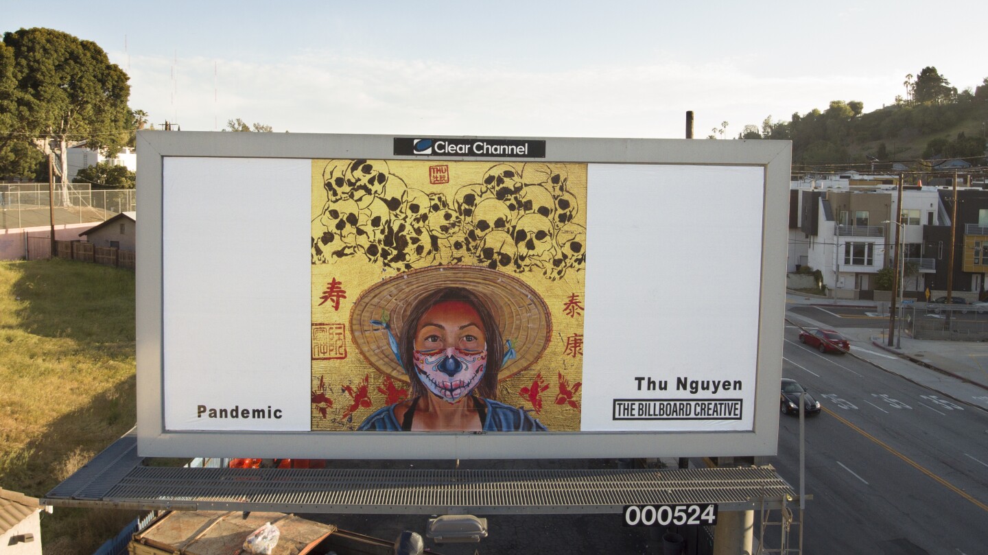 A billboard shows a portrait of a woman wearing a woven hat and a face mask. Behind her is a gold background and a pile of skulls drawn on.
