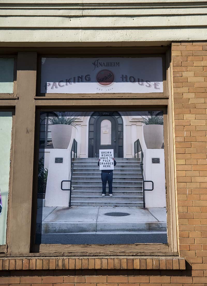 William Camargo holds up a sign that says, "On this side of the wall is a predominantly Brown working class neighborhood" as part of his exhibition, "Origins and Displacements, Vols. 1 & 2" at MUZEO | Michael Quintero