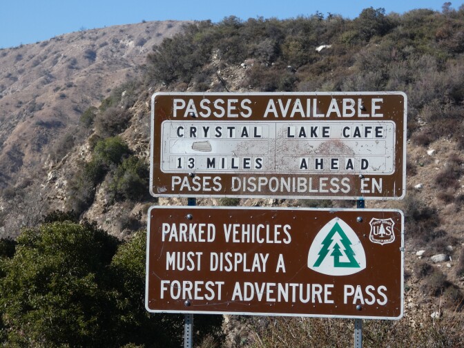 A metal, brown road sign reads: "Passes available: Crystal Lake Cafe, 12 miles ahead. Below, it also reads, "Parked vehicles must display a forest adventure pass."
