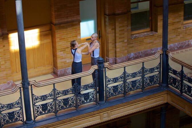 Two performers wearing black pants and suspenders perform on a balcony setting with ornate grills.