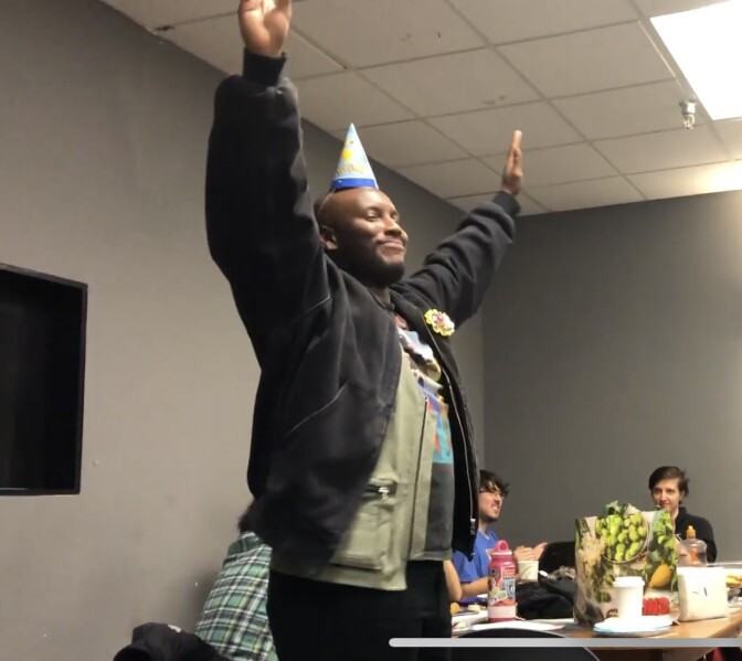 Marvin Bynoe stands with his hands raised above his head and a smile on his face. He's wearing a birthday hat. He's in a classroom setting, with classmates sitting behind him.