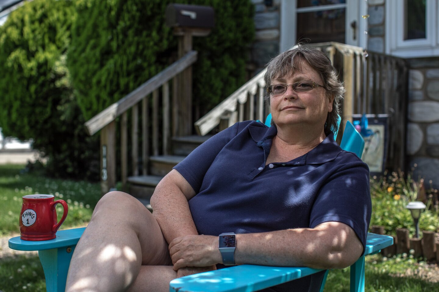 Susan sits on a bright blue wooden lawn chair. She's sitting leaned back, with her legs folded and her arms crossed. Patches of sunlight light different parts of her body as she smiles with a gaze slightly off from the camera.