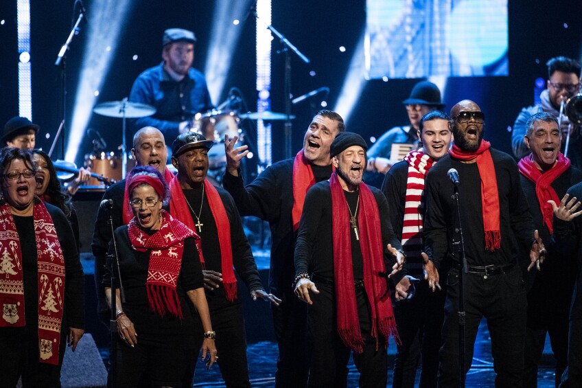 A group of men and women sing on stage, each wearing a black long sleeve shirt and black pants with a red scarf tied around their neck. Behind the group are other instrumentalists in the background including a drummer, accordion player, and trombone.