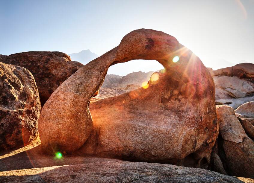A keyhole red rock formation in a desert. A sun shines behind the rock, sun rays stretching across the photo.