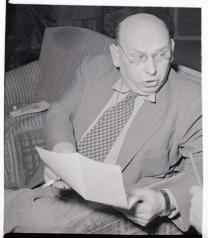 A black and white photo of Hanns Eisler reading a document. He wears a suit and tie and has his mouth open, halfway through. In one of his hands, a cigarette rests between his fingers.