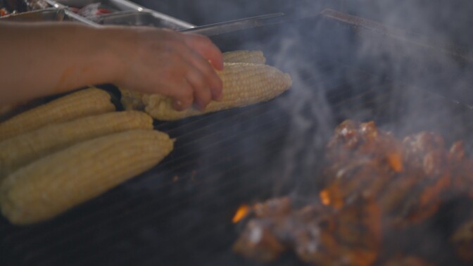 Golden yellow ears of corn sit on a flaming grill. Next to it are charred meats. Smoke rises from below the metal grill.