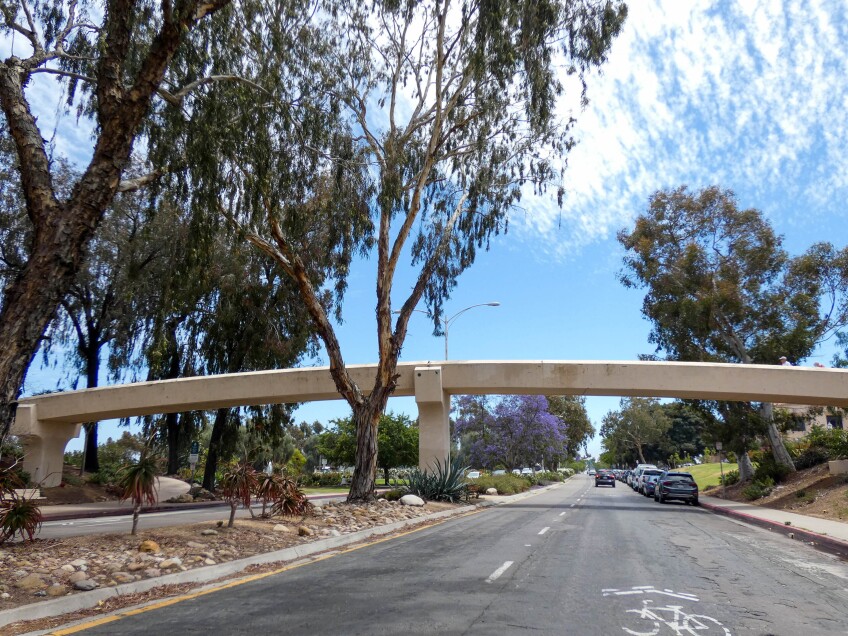 A concrete pedestrian foodbridge stretches over a four-lane road. Cars are seen parked along the curb on the right and one is driving down a lane. Large trees tower of the road and surrounding park area. 