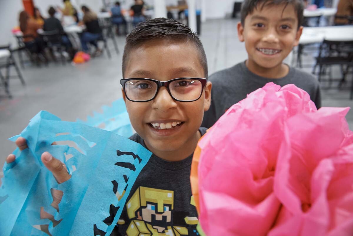 A young boy holds up blue papel picado and a pink paper flower at Self Help Graphics' Dia de los Muertos season of workshops, 2019. | Ken Marchionno