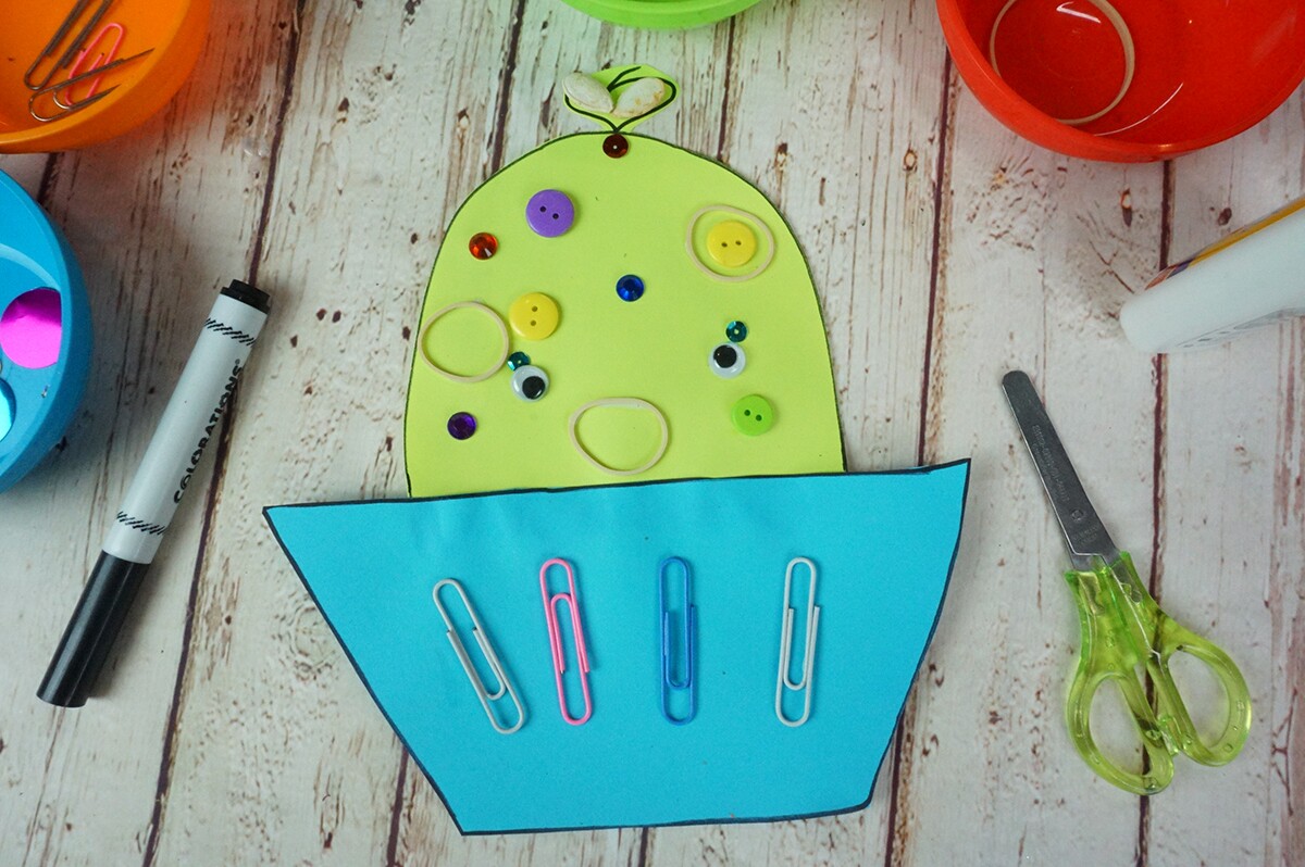 Un recorte de papel verde y azul con forma de pastelito decorado con ojos saltones, lentejuelas, clips de papel y ligas.