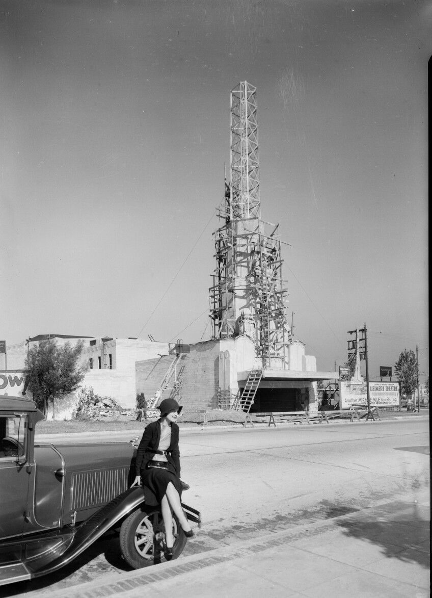 Photograph of woman sitting on automobile showing the Leimert Theater under construction in the background, Leimert Park, Los Angeles, CA, 1931.