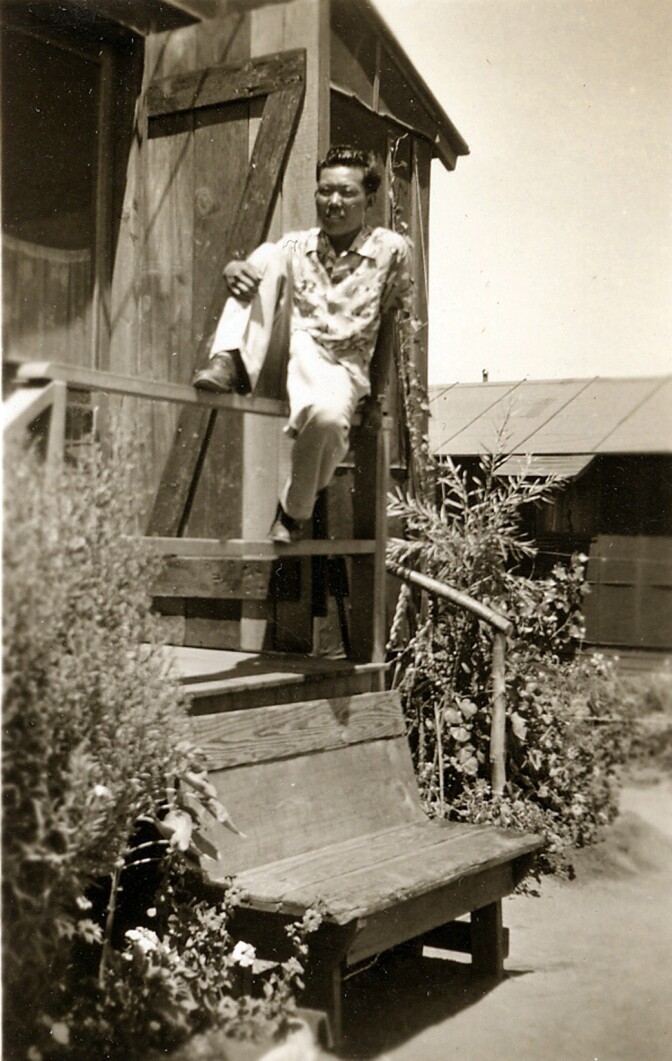 An old file photo of a young George Izumi sitting on a railing near a small wooden house. He is sitting on the railing with one leg up.