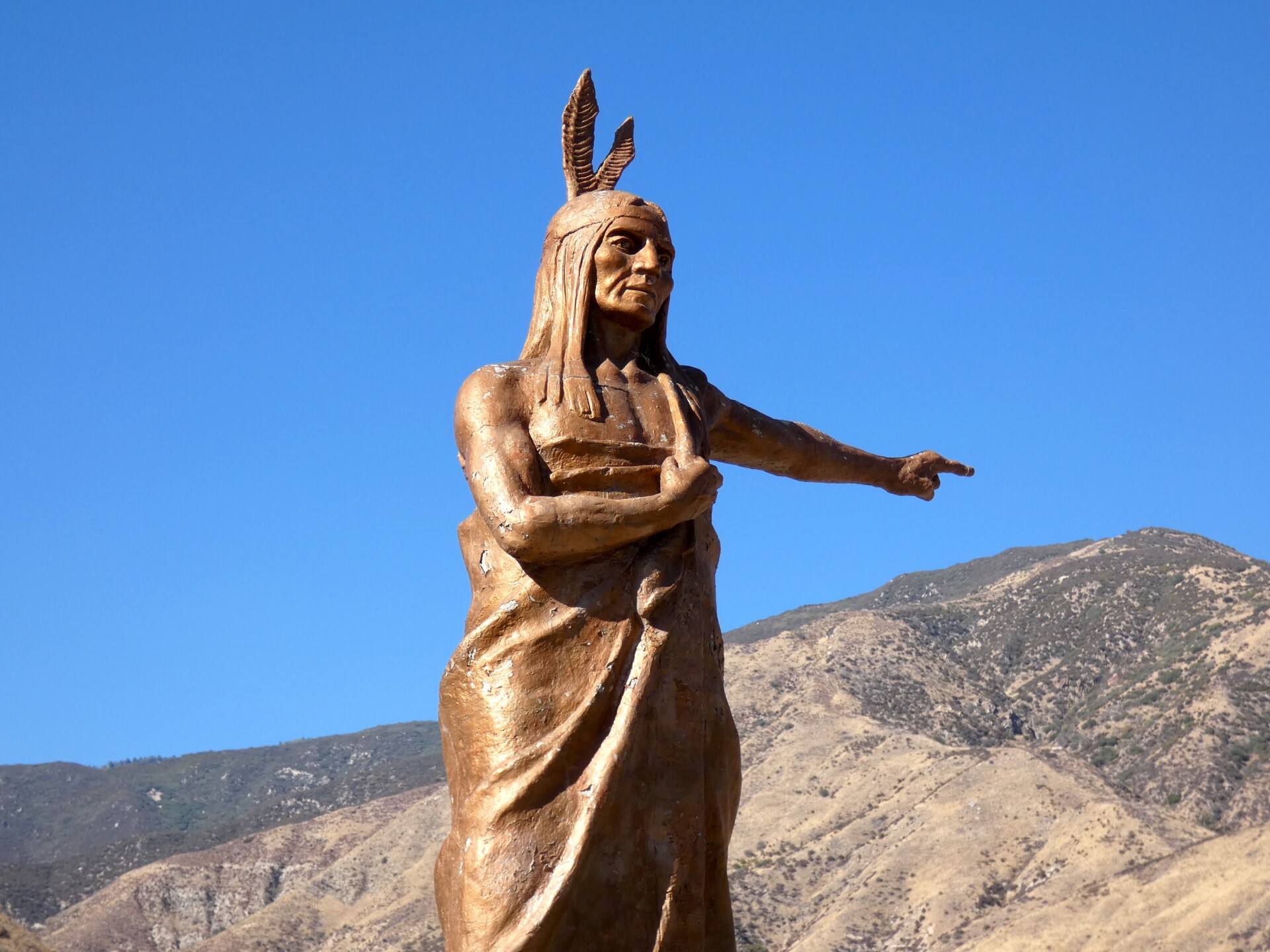 A bronze statue of a Native American points to the left. The Native American has shoulder length hair and has two feathers coming out from behind their head, attached to a headband.