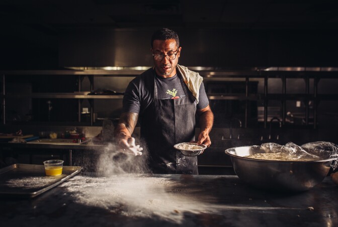 Chef Chris Williams stands over a metal kitchen counter and dusts flour over the surface from a small metal bowl in his other hand. He's wearing a black apron and has a kitchen cloth draped over his shoulder. A large metal bowl sits to his left with the plastic wrap partially covering the bowl's contents.