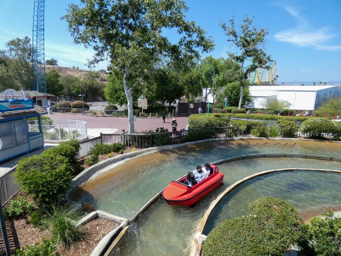 Three passengers on a red ride car sit directly behind each other. The ride car floats over a river stream that is paved by two, short concrete columns on either side.