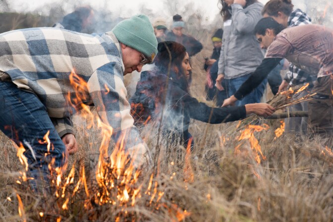Two individuals crouch in the tan brush, tending to the fire as others do the same in the background.