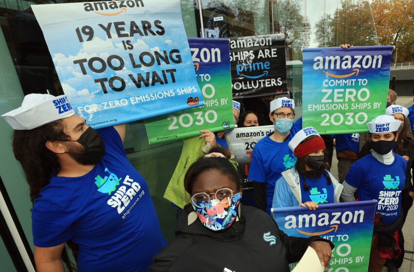 A group of masked protestors wearing blue T-shirts that read in white lettering "SHIP IT TO ZERO" hold signs with Amazon logos, reading "COMMIT TO ZERO EMISSIONS SHIPS BY 2030" and "19 YEARS IS TOO LONG TO WAIT"