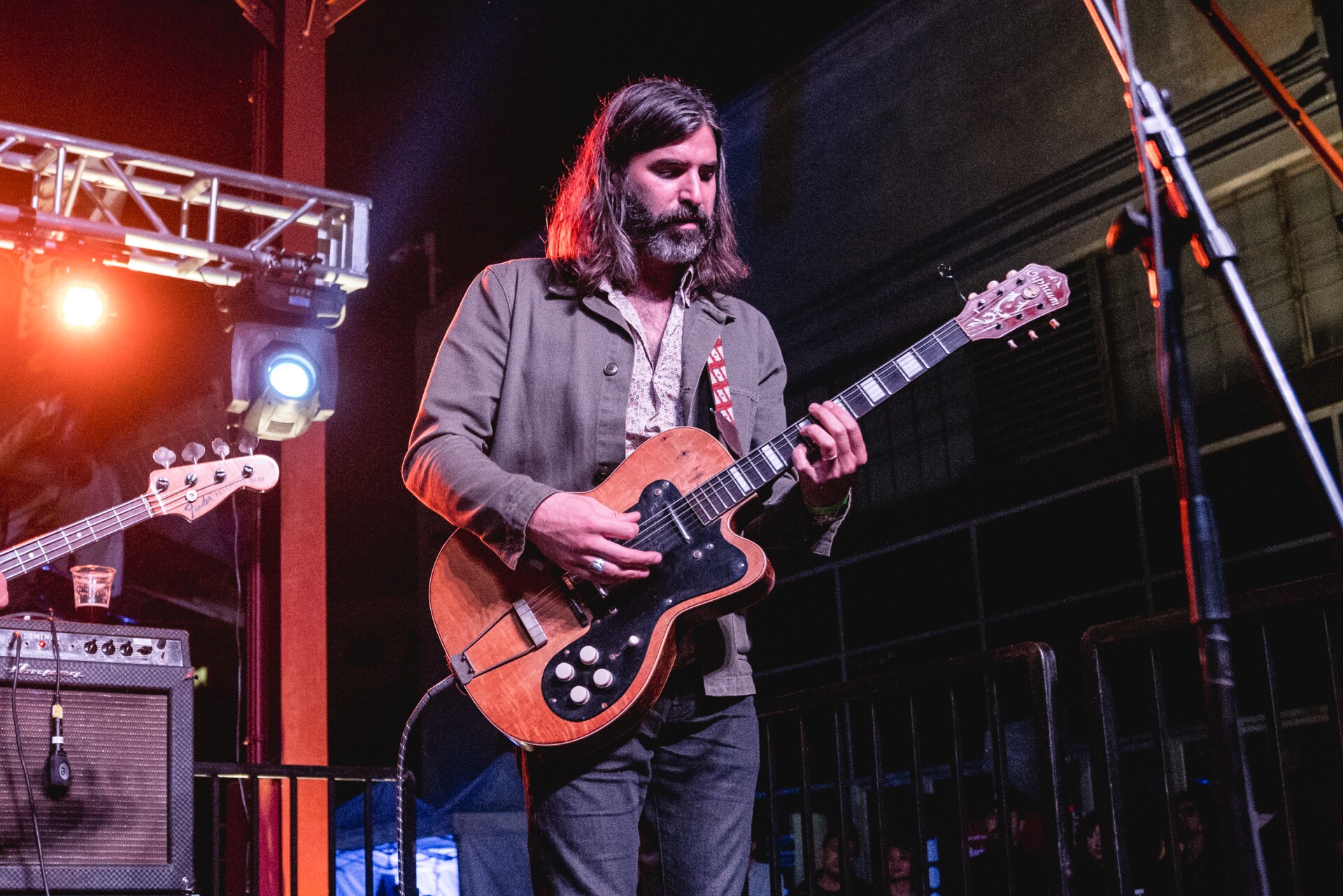 A man plays an orange electric guitar on stage. He's wearing a gray suit jacket. His hair is long, about shoulder length, and he has a short beard with specks of gray in it.