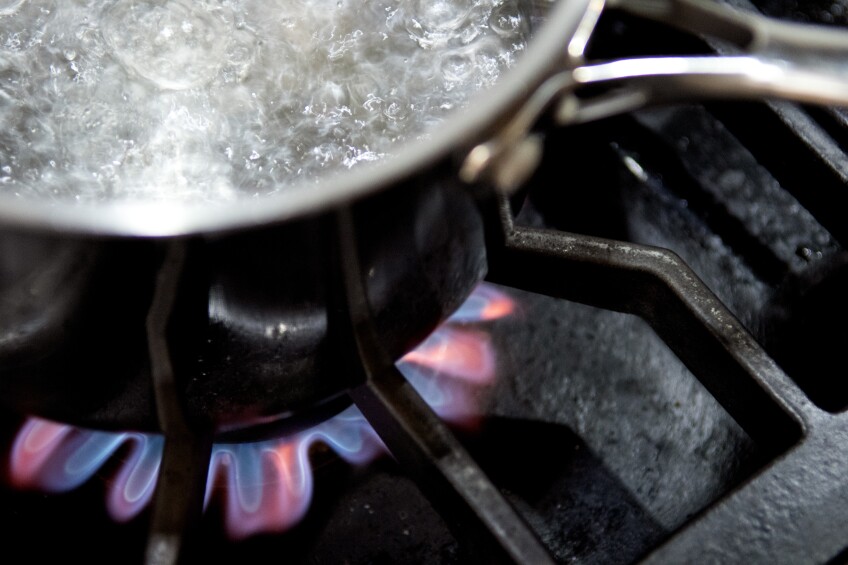 A stainless steel pot containing boiling water sits on top of a lit burner of a gas-powered stovetop