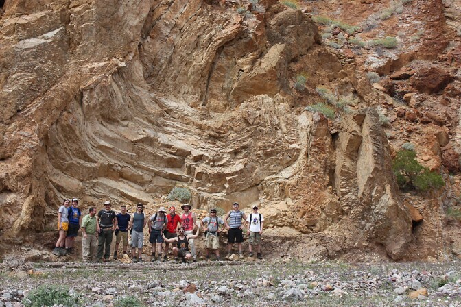 A group of students in hiking gear pose in front of an orange-brown rock formation with jagged edges. A man is kneeling in front of the group of young adults. The man kneeling is geologist Sammy Castonguay. Everyone is wearing hiking gear/athletic wear.