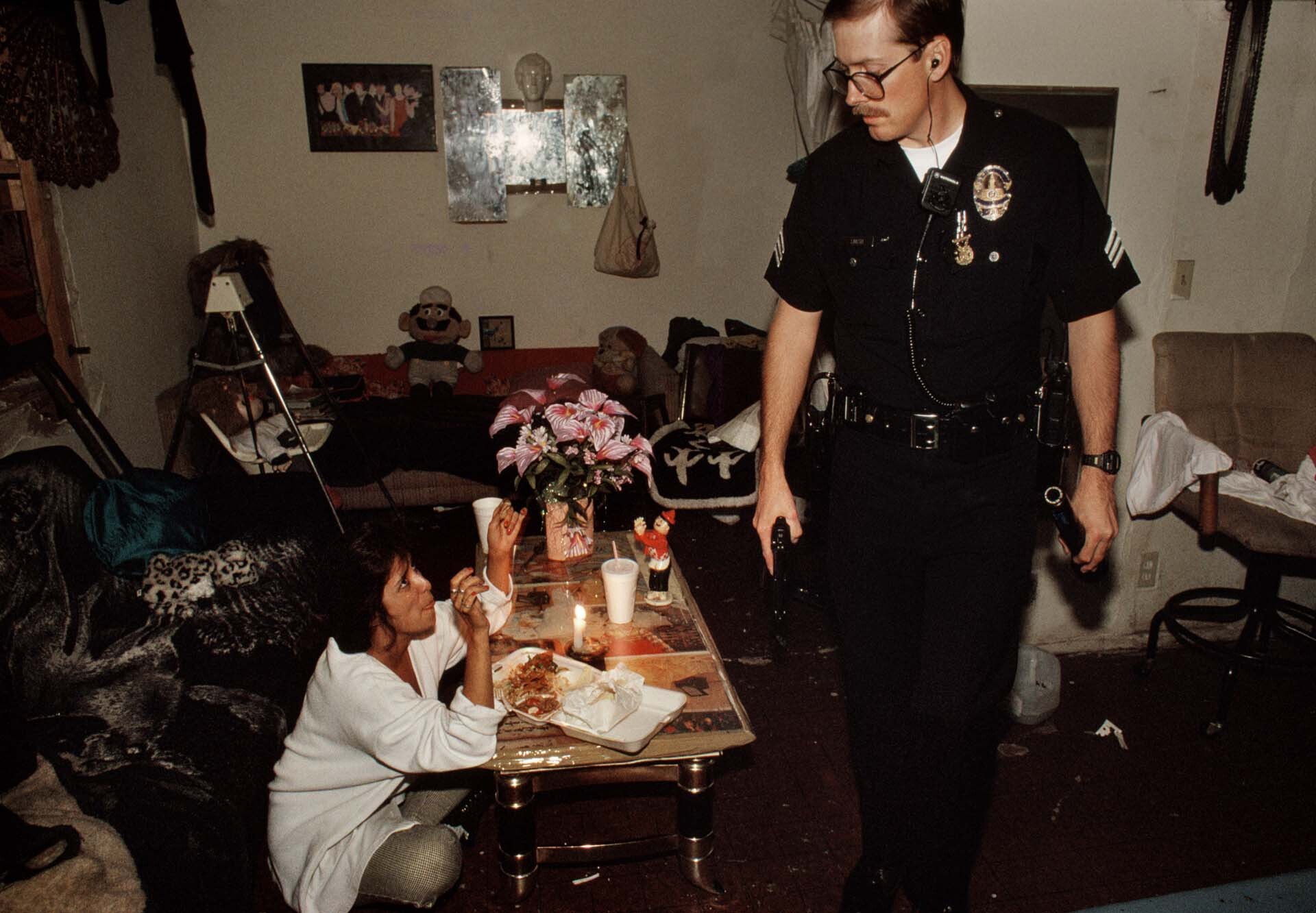 A woman in an abandoned motel where she has been living continues to eat dinner as police search the building for a murder suspect. | Joseph Rodriguez