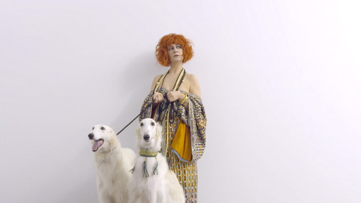 Still from Border Blaster 506, "Rhythmic Rituals:" A woman stands in front of a white hall with two white dogs on leashes.