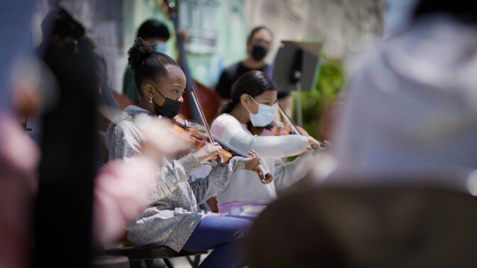 Two young girls are seated next to each other, playing the violin in the open air in the middle of an orchestra. They both wear face masks. They are framed by two people seated in the foreground who are very blurred.