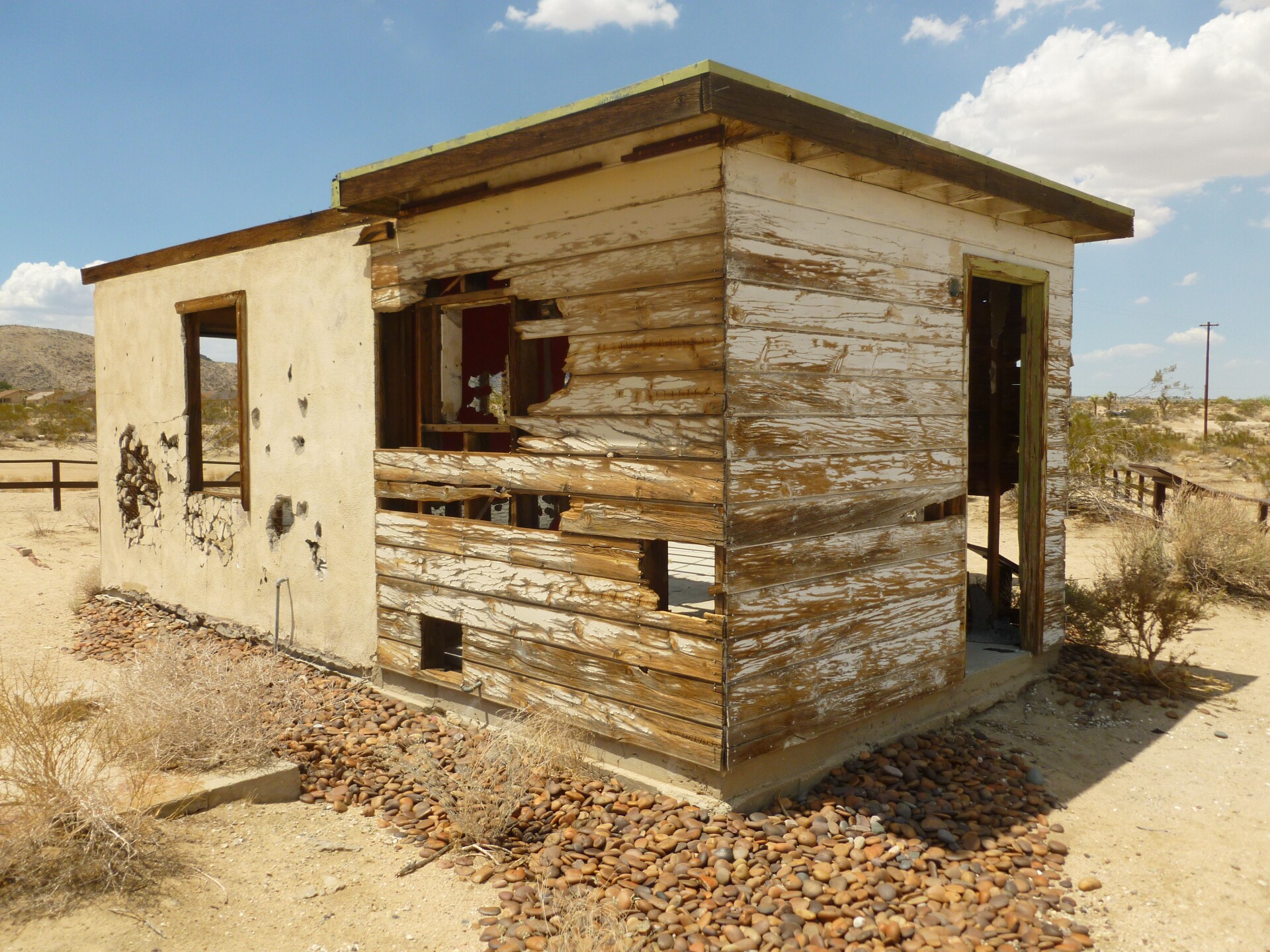 A small, abandoned and destroyed desert homestead made out of wood and concrete. The wood horizontal panels are worn out and parts of the wall are entirely gone. The concrete walls are crumbling, with pieces of it missing from the face.