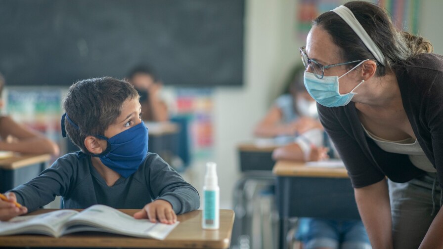 A masked teacher helps a small masked child. istock