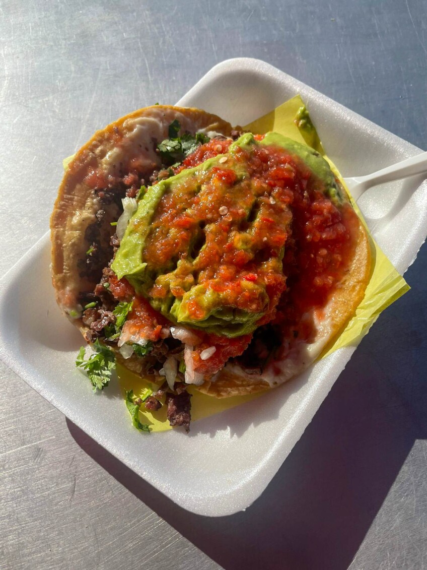 A taco filled with carne asada topped with chunky green guacamole, runny raw tomato salsa, white onions and cilantro. The taco sits on top of a styrofoam rectangular plate.